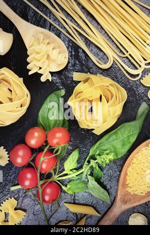 Italienische Pasta auf schwarzem Hintergrund. Spaghetti, pappardelle, orzo, farfalle und andere Arten, mit Tomaten und Basilikum Stockfoto