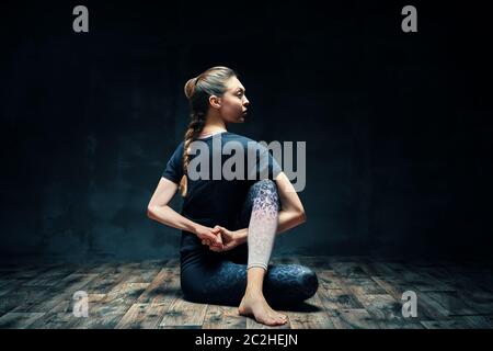 Junge schöne Frau, die Yoga Asana halb herr der Fische Pose auf dunklen Raum Stockfoto