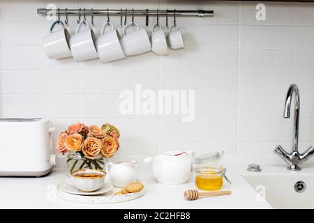 Hochzeitsstrauß aus rosa und orange Lisianthus, antirrhinum und Eukalyptus in Glasvase auf Küchentisch. Schwarzer Tee in einer weißen Tasse, Honigglas und Löffel Stockfoto