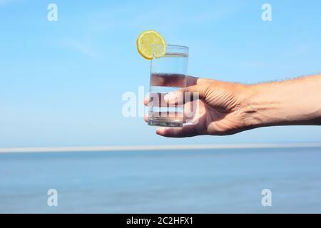 Eine Hand hält ein volles Glas Wasser vor einem verschwommenen Hintergrund mit Meer und Himmel Stockfoto