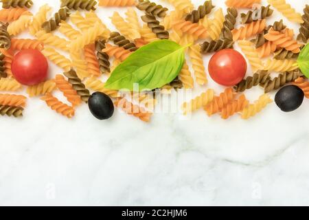 Italienische Küche Hintergrund. Fusilli-Nudeln mit schwarzen Oliven, Kirschtomaten und einem Basilikumblatt, von oben auf weißem Marmor geschossen Stockfoto