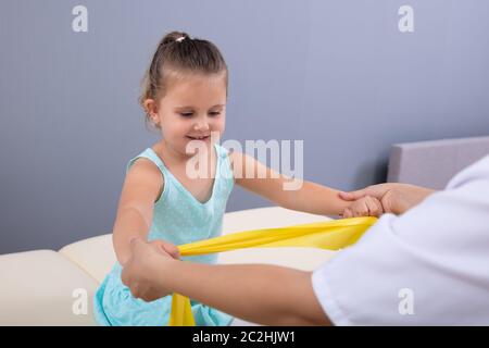 Weiblichen Physiotherapeuten Helfen süsse kleine Mädchen tun Übung mit Gummizug in physikalische Therapie Klinik Stockfoto