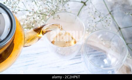 Gießen schwarzen Tee aus Glas Teekanne in Doppel-Wandbecher. Goldener Tee. Leere Becher. Heller weißer Holztisch. Stockfoto