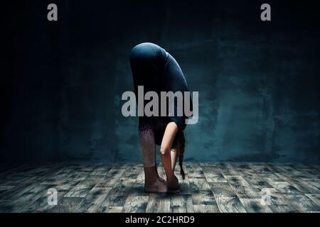 Junge Frau, die Yoga tut, die nach vorne gebeugt im dunklen Raum posiert Stockfoto