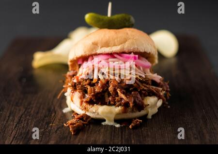 Langsam gekochtes Pulled Schweinefleisch mit BBQ-Sauce, eingelegte Zwiebeln, Krautsalben und Käse in einem Hamburger-Brötchen mit Dill Pickle und Chips als Beilagen serviert. Es ist chaotisch. Stockfoto