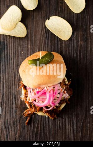 Langsam gekochtes Pulled Schweinefleisch mit BBQ-Sauce, eingelegte Zwiebeln, Krautsalben und Käse in einem Hamburger-Brötchen mit Dill Pickle und Chips als Beilagen serviert. Es ist ein Chaos Stockfoto