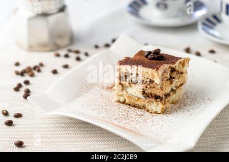 Köstliche hausgemachte Tiramisu Kuchen Scheibe auf einem weißen Teller. Ein klassisches italienisches Dessert. Im Hintergrund Espresso Kaffeetassen und verschüttete Kaffeebohnen. Stockfoto