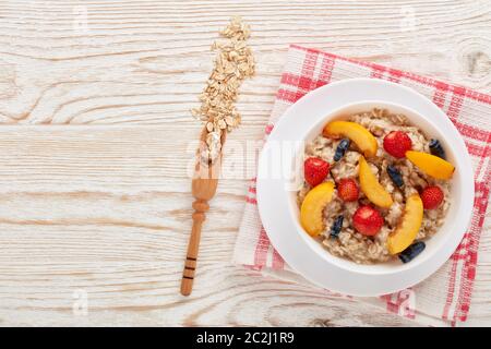 Haferbrei Haferbrei mit frischen Beeren und Obst in weißer Schüssel auf hellem Holzhintergrund. Draufsicht mit Kopierbereich. Konzept der gesunden Lebensweise, Gesundheit Stockfoto