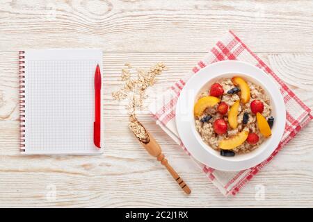 Haferbrei Haferbrei mit frischen Beeren und Obst in weißer Schüssel, leeres Notizbuch auf hellem Holzhintergrund. Draufsicht. Konzept der gesunden Lebensweise, gesund Stockfoto