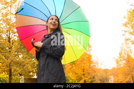 Ziemlich schöne junge Frau unter Rainbow Regen Regenschirm in den Park auf der Seite betrachtet. Stockfoto
