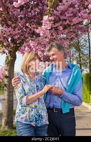 Liebevolles Senioren-Paar im Frühling posiert mit Händen halten Blick liebevoll auf einander unter einem Baum mit rosa Blüten bedeckt. Stockfoto