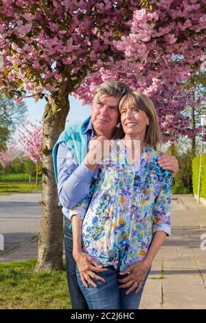 Liebevolles älteres Paar, das den Frühlingssonne genießt, steht in einer engen Umarmung mit Gesichtern zur Sonne vor der bunten rosa Blüte auf einem Baum. Stockfoto