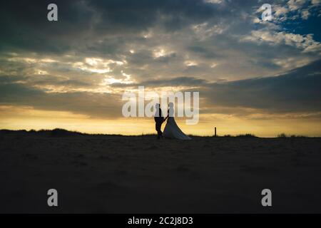 Silhouette von frisch verheiratetetes Paar am Abend Strand. Braut und Bräutigam gehen am Horizont. Dramatischer Himmel mit Sonnenstrahlen zwischen Wolken im Hintergrund. Glück, Liebe Stockfoto