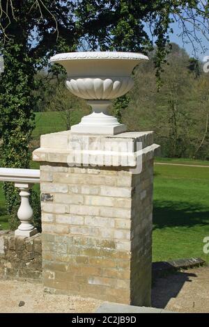 Ornamental jardiniere auf einer Ziegelsäule, die an einer alten Wand und Balustrade aus hellgrauen Ziegelsteinen befestigt ist. Rasen, Bäume und blauer Himmel Stockfoto