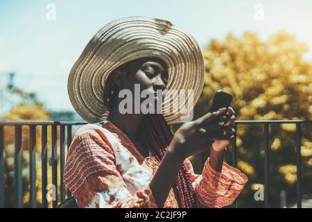 Ein charmantes schwarzes Touristenmädchen in Strohhut und Sundress, das draußen auf einem Balkon sitzt und ihr Handy benutzt und einen warmen sonnigen Tag genießt; afrikanisch Stockfoto