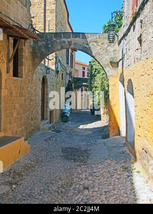 Typische ruhige gepflasterte Straße in rhodos Stadt mit alten gelb bemalten Häusern und einem Steinbogen mit sonnenbeschienenen Sommerhimmel Stockfoto