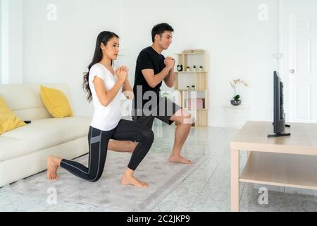 Junge asiatische Paar tut Kniebeugen Training zusammen und suchen TV zu Hause, Mann und Frau arbeiten zusammen im Wohnzimmer stehen, fit Paar führen Stockfoto