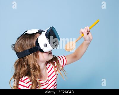 Lächelndes positives Mädchen trägt Virtual-Reality-Brille Brille Headset singt mit Kochlöffel, neue Generation, Konzept vor blauem Hintergrund Stockfoto