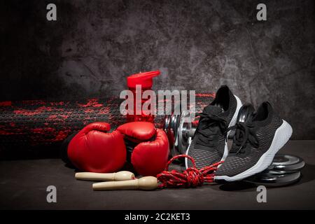 Sportausrüstung. Boxhandschuhe, Schaumstoffrolle, Hantel, Springseil, Wasserflasche und Sneaker auf dunkelgrauem Hintergrund mit Kopierfläche. Fitness, Sport Stockfoto