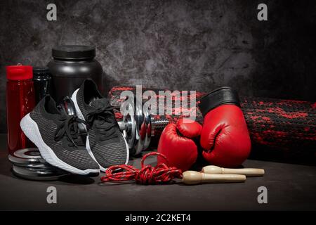 Sportnahrung, Boxhandschuhe, Schaumstoffrolle, Hantel, Springseil, Wasserflasche und Sneaker auf dunkelgrauem Hintergrund mit Kopierfläche. Fitness, Sport Stockfoto