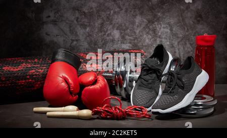 Sportausrüstung. Boxhandschuhe, Schaumstoffrolle, Hantel, Springseil, Wasserflasche und Sneaker auf dunkelgrauem Hintergrund mit Kopierfläche. Fitness, Sport Stockfoto