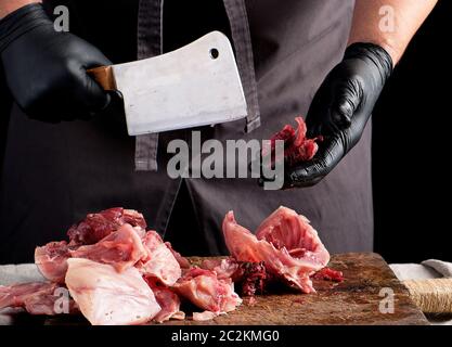 Koch in Schwarzem latex Chunks in Stücke schneidet rohes Fleisch auf einem braunen Holzbrett, kochen auf einem dunklen Hintergrund Stockfoto