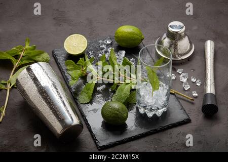 Set von Bar-Tools: Cocktail Shaker, Muddler und Glas mit Eis, Limette und Minze auf dunklen Stein Tisch. Draufsicht. Werkzeug des Barkeepers. Mojito Cocktailzubereitung Stockfoto