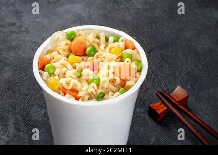 Ramen cup close-up, instant Soba Nudeln in eine Schale aus Kunststoff mit Gemüse, auf einem schwarzen Hintergrund mit Stäbchen Stockfoto