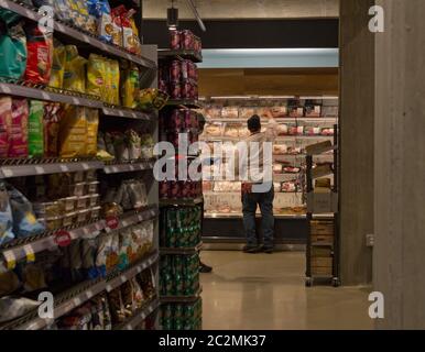 Baltimore, Maryland, USA. Juni 2020. Rückansicht des männlichen Mitarbeiters im Arbeitsschürze und Baseballmütze/Hut am umgesiedelten Baltimore Harbor East Whole Foods Market in der South Central Avenue 711, der am Eröffnungstag Pakete mit Hühnchen im Kühlbereich im Laden stapelte, mit Regalen mit Chips/Chips und sechs Dosen mit Soda Pop/Soft Getränke im Gang im Vordergrund. Der frühere Standort dieser Niederlassung der multinationalen Supermarktkette im Besitz von Amazon, die einige Tage zuvor geschlossen hatte, befand sich in der nahe gelegenen Fleet Street. Kay Howell/Alamy Live News Stockfoto