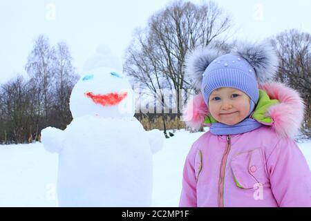 Porträt eines lächelnden Babys in amüsanten Wintermütze mit zwei lustigen Ohren. Kleines Mädchen und Schneemann Kopf Stockfoto