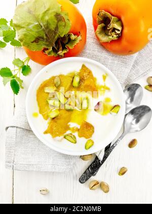 Dessert aus Joghurt, Persifon und Honig mit Vanille, Kardamom und Pistazien in einer Schüssel auf Serviette, Minze und Löffel auf dem Hintergrund von hellem Holz B. Stockfoto