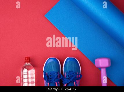 Zwei blaue Turnschuhe mit Schnürsenkeln, einer Flasche Wasser und einer blau verdrehten Neopren-Matte auf rotem Texturhintergrund, Draufsicht, Sportset Stockfoto