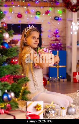 Das zehnjährige Mädchen sitzt am Weihnachtsbaum und umarmt ein Geschenk Stockfoto