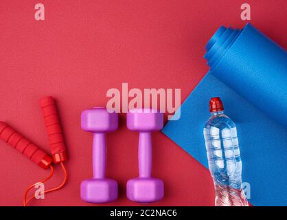 Zwei violette Kunststoff-Hanteln, eine Flasche Wasser auf einer roten Neopren-Matte, Draufsicht, Trainingsausrüstung Stockfoto