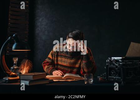 Bärtige Autor in Gläsern ein Buch zu lesen. Retro-Schreibmaschine, Feder, Kristallkaraffe, Bücher und Vintage Lampe auf dem Schreibtisch Stockfoto