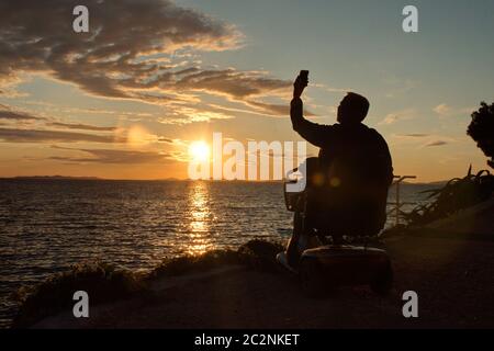 Behinderter älterer Mann, der im Rollstuhl sitzt und während des Sonnenuntergangs an der Adriaküste mit dem Handy telefoniert Stockfoto