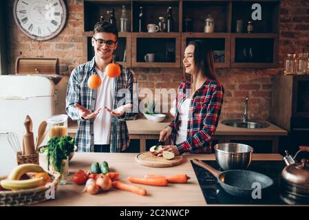 Junge Mann jongliert Orangen in der Küche, während Frau kochen Obst Frühstück. Mann und Frau Vorbereitung Gemüsesalat, glückliche Familie zusammen Stockfoto