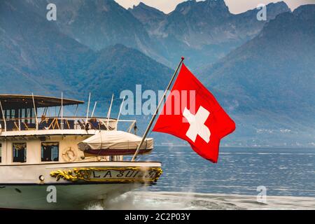 Der majestätische Raddampfer (La Suisse) gleitet über den Genfer See mit der Schweizer Flagge, die vor der atemberaubenden Bergkulisse winkt Stockfoto