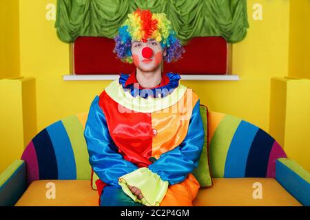 Clown in Regenbogenfarben Kostüm mit Make-up und roter Nase auf buntem Sofa sitzend. Stockfoto
