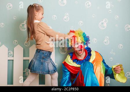 Kleines Mädchen zieht Clownhaare. Weiße Zaundekoration auf dem Hintergrund. Stockfoto