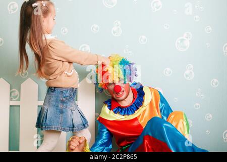 Kleines Mädchen zieht Clownhaare. Weiße Zaundekoration auf dem Hintergrund. Stockfoto