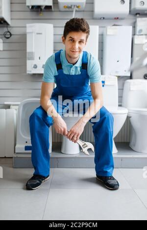 Klempner in Uniform sitzt auf der Toilette bei der Vitrine in Klempnerladen. Mann kauft Sanitär-Engineering-Werkzeuge und Geräte im Geschäft Stockfoto