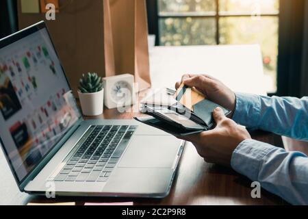 Asiatische Mann Hand zieht eine Kreditkarte aus seiner Tasche, um Online-Shopping auf dem Computer zu vervollständigen. Stockfoto