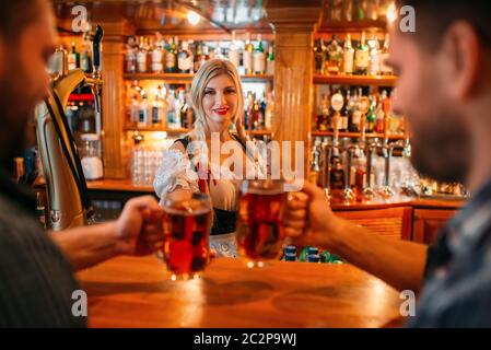 Zwei männliche Freunde klirren Becher mit einem Bier in der Kneipe, Kellnerin in der Zähler auf Hintergrund, oktoberfest Urlaub. Männer mit Brille Spaß in bar, barm Stockfoto