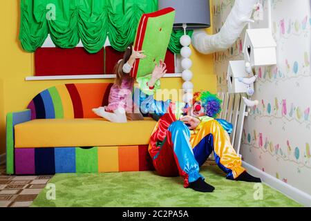 Glückliches kleines Mädchen schlägt Clown mit einem großen Kissen. Spielzimmer mit buntem Sofa im Hintergrund. Stockfoto