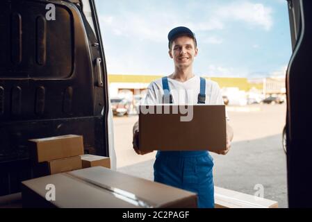 Liefermann in Uniform entlädt das Auto mit Paketen, Lieferservice. Mann steht an Pappverpackungen im Fahrzeug, männliche liefern, Kurier oder shippin Stockfoto