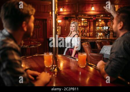 Freunde klirren Becher in Bier Bar, Kellnerin in der Zähler auf Hintergrund, oktoberfest Urlaub. Männer mit Brille Spaß im Pub, bardame in Tradit Stockfoto