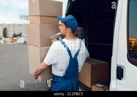 Postbote in Uniform entlädt das Auto mit Paketen, Lieferservice. Mann steht an Pappverpackungen im Fahrzeug, männlich liefern, Kurier oder Versand jo Stockfoto