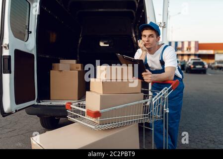 Deliveryman in Uniform hält Wagen mit Boxen am Auto mit Paketen, Lieferservice. Mann steht an Pappverpackungen im Fahrzeug, Mann liefern, c Stockfoto