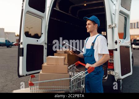 Deliveryman in Uniform hält Wagen mit Boxen am Auto mit Paketen, Lieferservice. Mann steht an Pappverpackungen im Fahrzeug, Mann liefern, c Stockfoto